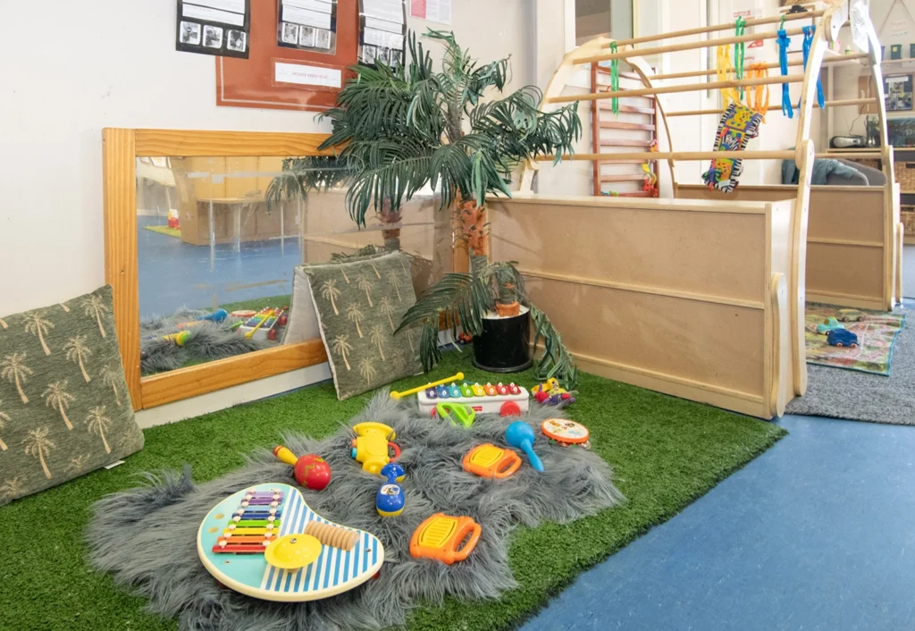 A children’s play area in the kindergarten features a mirror, cushions, a fake palm tree, and various colorful musical toys on a gray rug over green turf. A small wooden climbing structure is in the background, creating an ideal early learning environment.