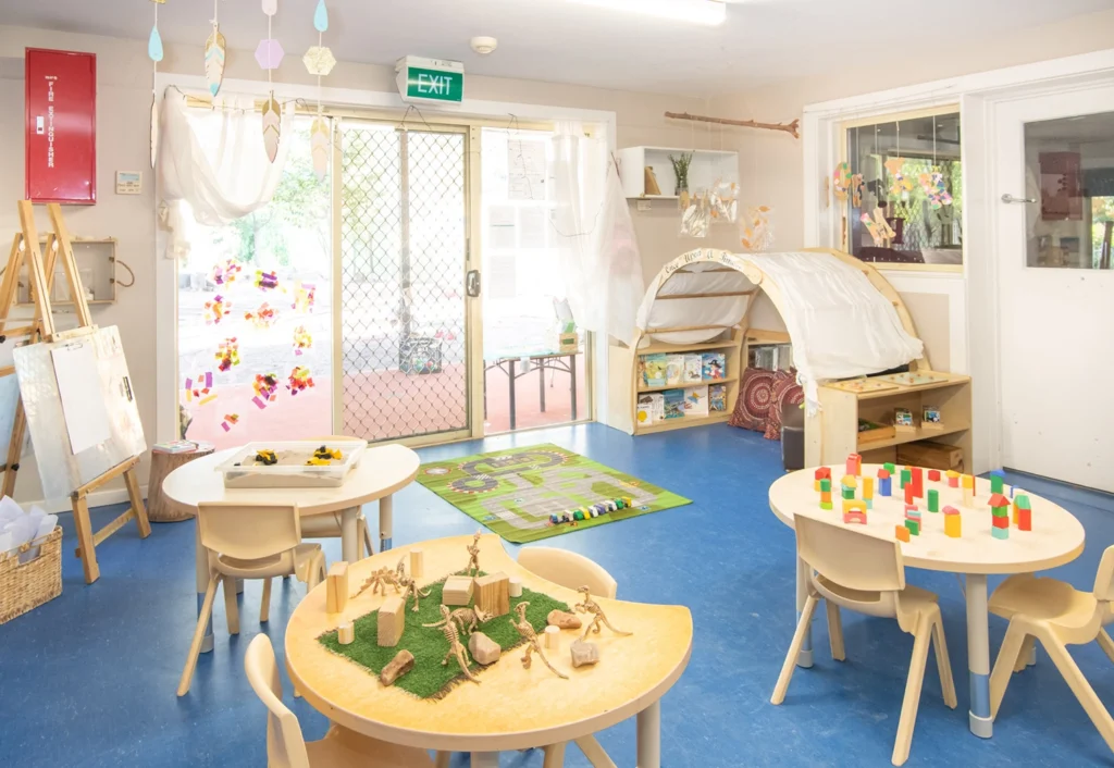 A well-organized kindergarten classroom with small tables, chairs, educational toys, and a play area rug. Large windows provide natural light, and art supplies are visible in the background, fostering an ideal environment for early learning.