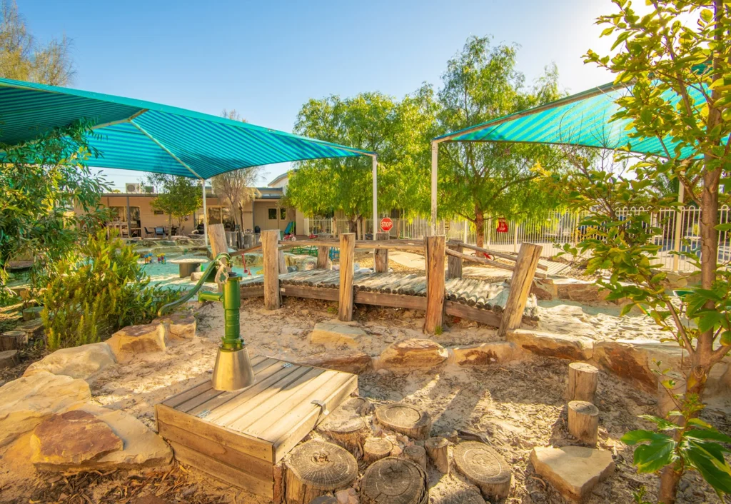 A sunny outdoor play area at the preschool features shaded canopies, wooden structures, a water pump, and natural elements like trees and rocks.