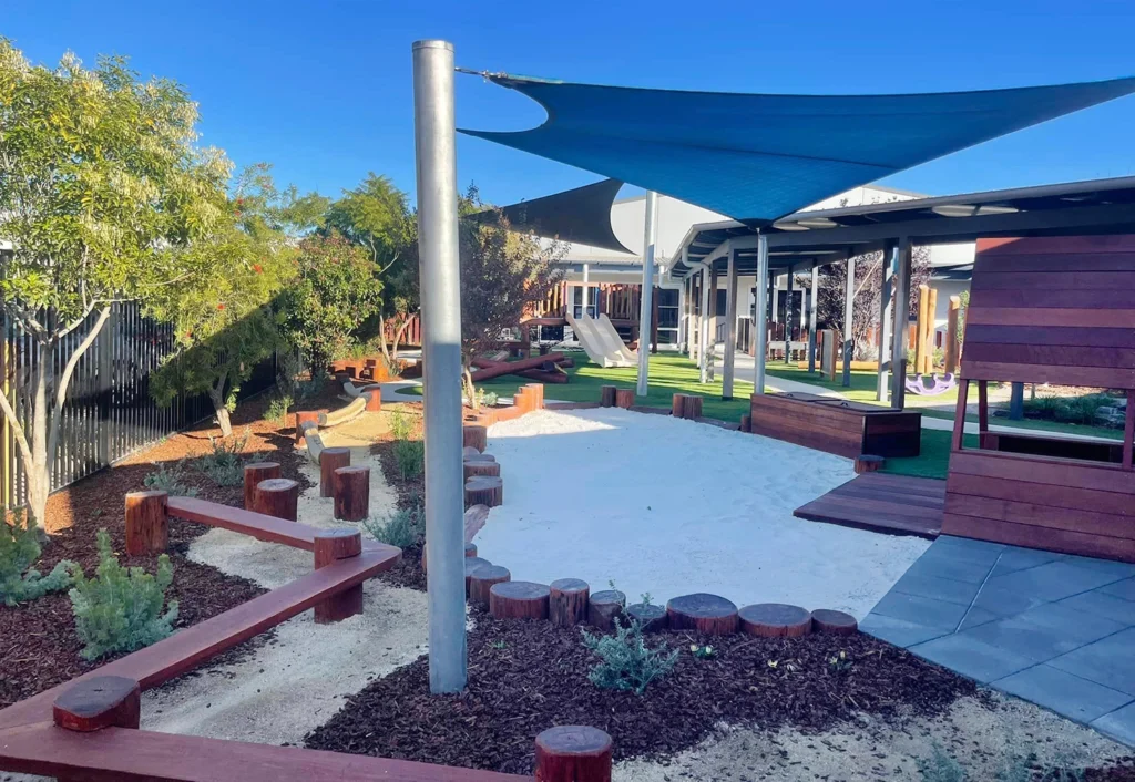 A playground area with wooden climbing structures, a shade sail, and sandpit. Trees and small plants are scattered throughout, creating a perfect environment for preschool fun. Buildings are visible in the background, suggesting this could be part of a day care or childcare facility.