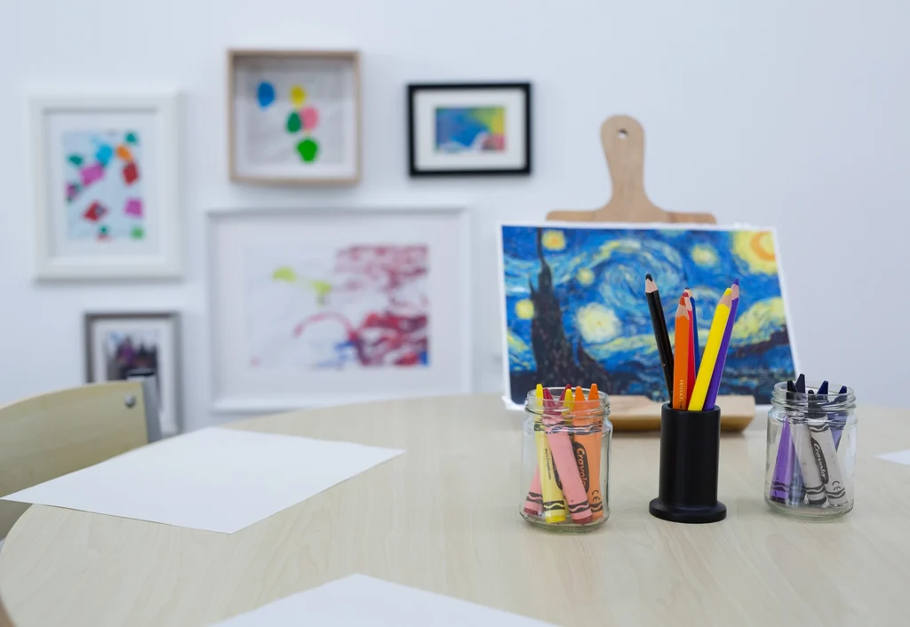 A round table with blank white papers, jars of colored pencils, and crayons sits in the vibrant art corner. A small painting and framed artworks are visible on the wall in the background. The setting appears to be an engaging preschool classroom.