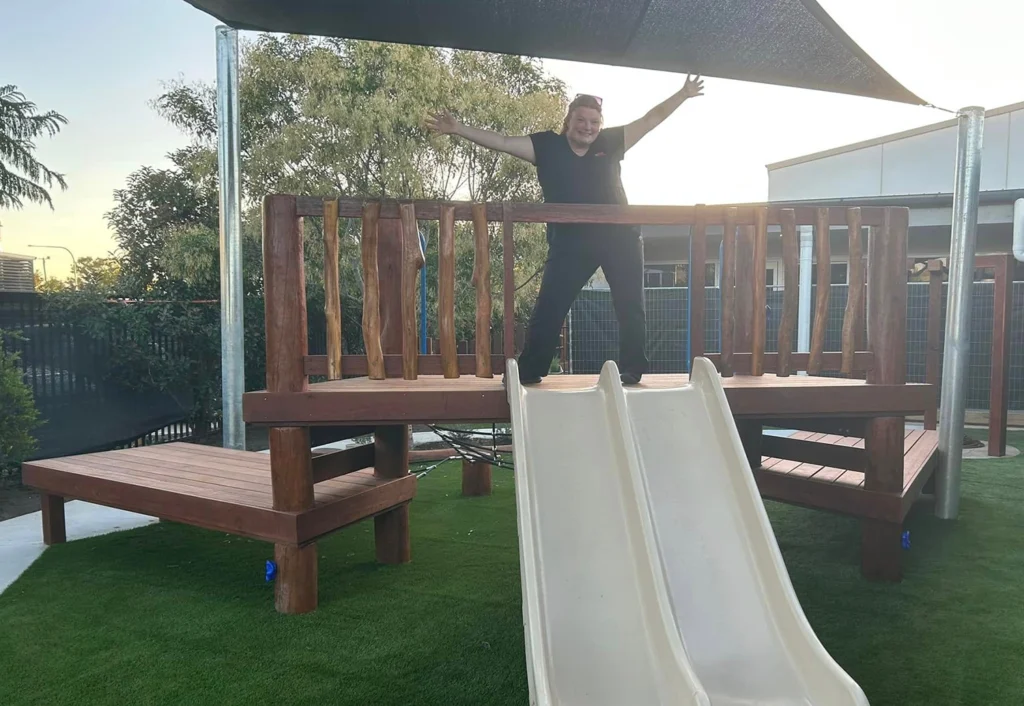 A person stands with arms outstretched on a wooden platform featuring a slide, at a kindergarten playground with artificial grass and partial shade coverage.