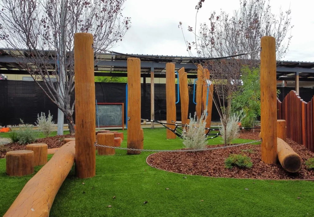 A playground designed for kindergarten with wooden balance beams, vertical poles, and a hammock stands on a grassy area with mulch and young trees.