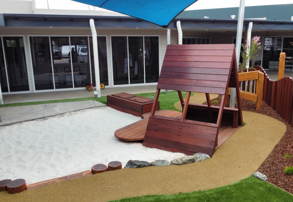 Outdoor playground area with a wooden play structure, a sandpit, artificial grass, and a shade sail above—ideal for preschool adventures. A building with large windows in the background enhances the bright and welcoming early learning environment.