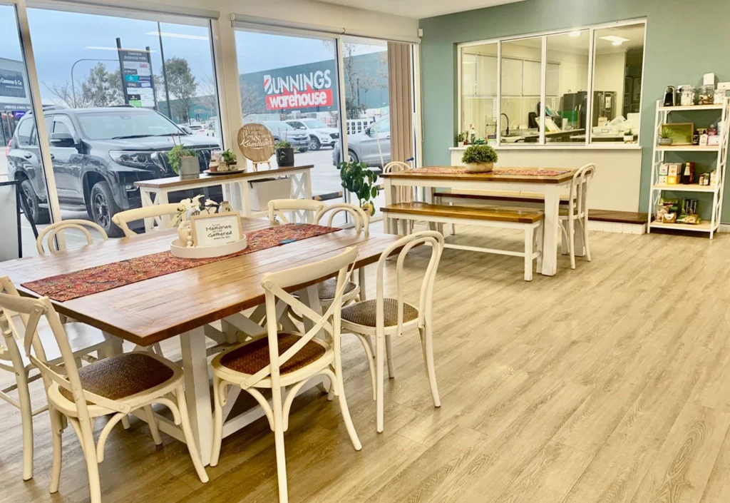 A well-lit cafe with wooden tables and white chairs, a view of the Bunnings Warehouse store through large windows, and a small kitchenette area with shelves and a refrigerator in the background – perfect for preschool parents to relax while their little ones enjoy early learning activities nearby.