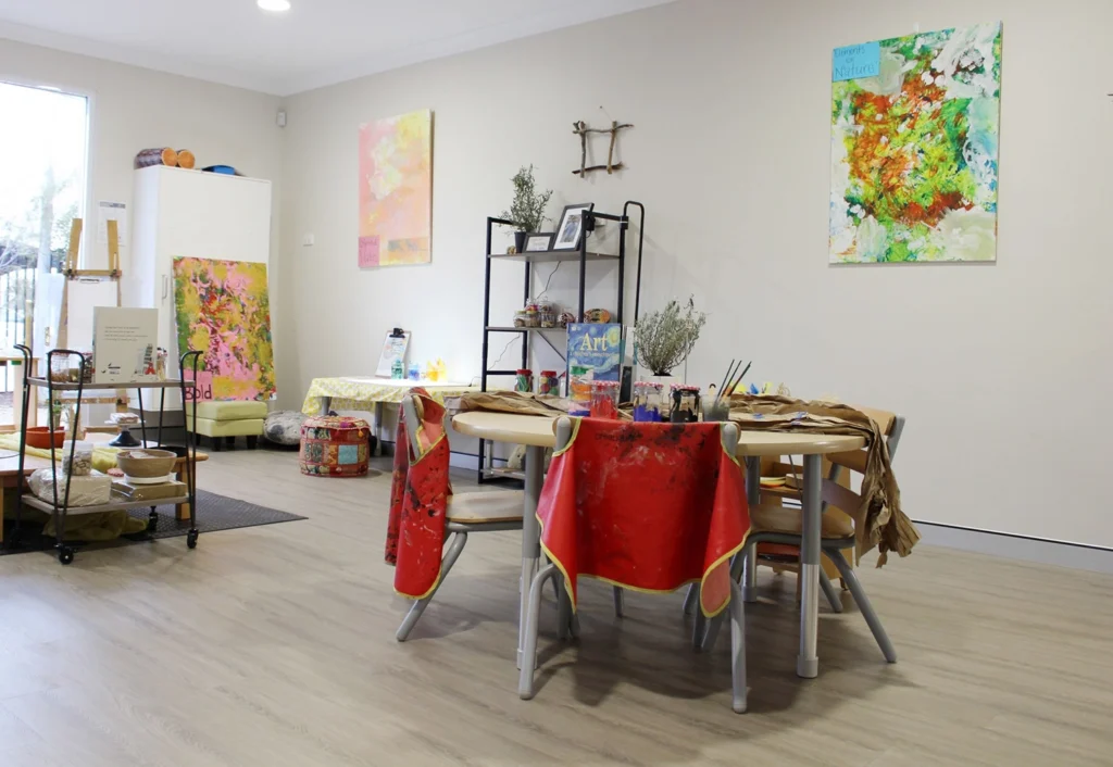 A small art studio designed for preschool children, with their paintings adorning the walls, a round table with art supplies and red-paint-stained tablecloths, and shelves filled with additional supplies and decorations. This vibrant space fosters creativity in an early learning environment.