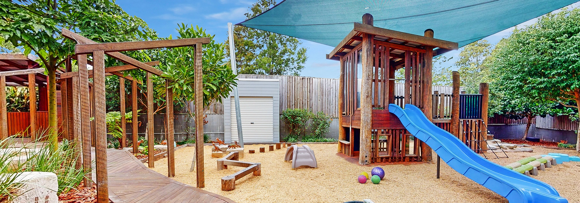 A children's outdoor playground with wooden play structures, a blue slide, various climbing equipment, and colorful play balls on a sandy surface offers endless fun at this preschool. There’s also a shaded area for breaks and trees along with a fenced boundary in the background.