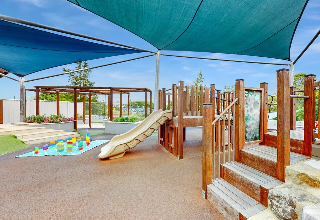 Playground with wooden structures, a slide, and a shaded area. Colorful toys and a blanket are laid out on the ground, perfect for a preschool outing.