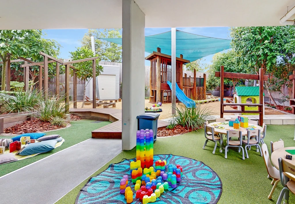 An outdoor playground at the day care with a slide, climbing structures, and shaded areas. Various toys and activity stations are set up, including a table with colorful stacking toys and seating areas, perfect for early learning or kindergarten adventures.