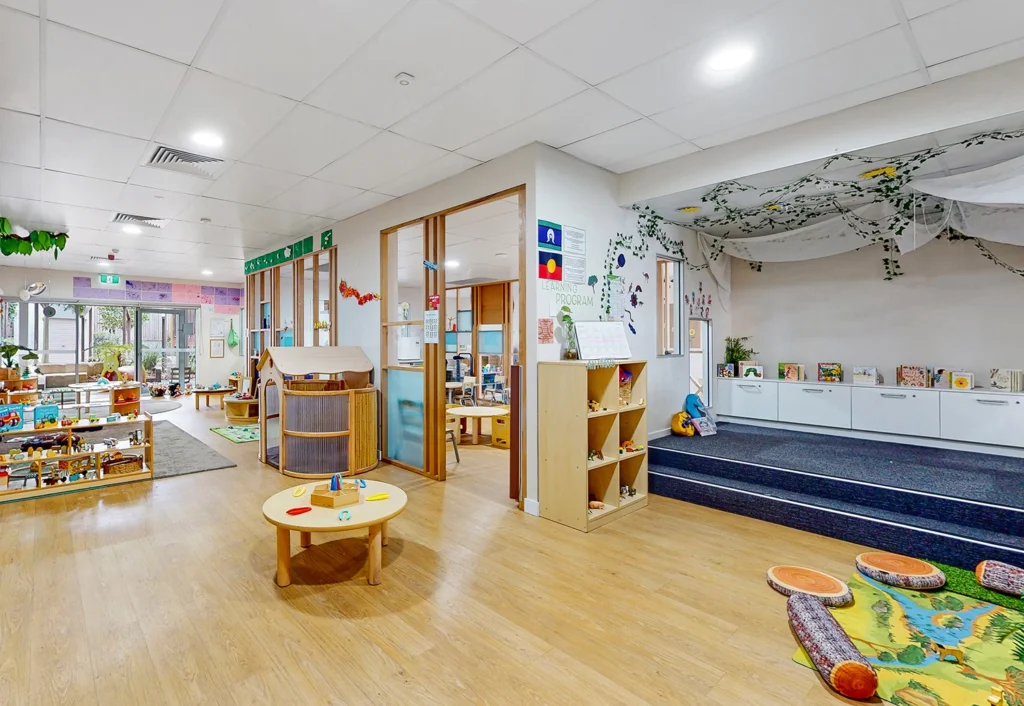 Spacious and brightly lit kindergarten classroom with educational materials, a platform area, wooden furniture, and children's play items. Ceiling decor includes hanging plants and paper cutouts.