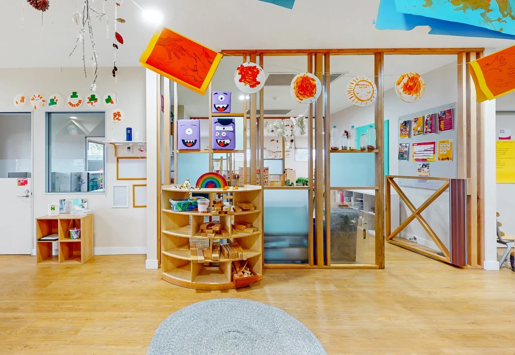 A brightly decorated kindergarten classroom with wooden shelves, art supplies, colorful artwork on walls, and a circular rug on the floor. Various crafts and educational materials are displayed throughout the room, creating a cheerful environment perfect for early childhood education.