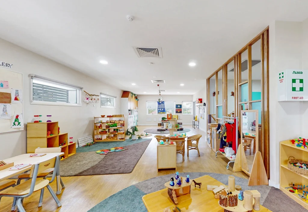 A well-lit preschool classroom with various play areas, children’s artwork on the walls, wooden furniture, and organized toys. A first aid kit is mounted on the wall, ensuring the safety of everyone in this nurturing childcare environment.