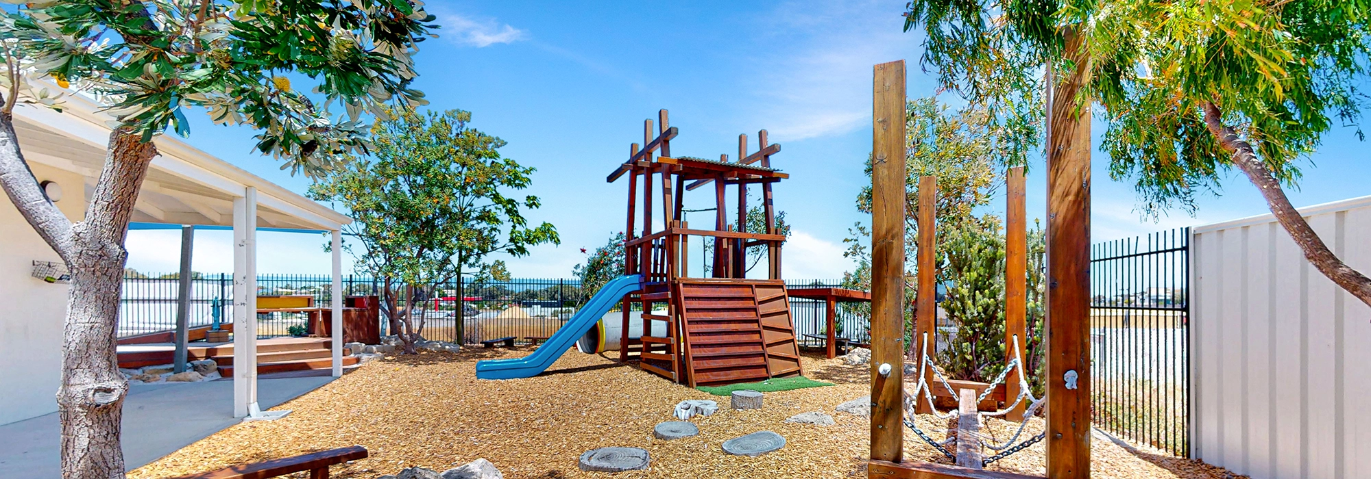 Outdoor playground with a wooden climbing structure, blue slide, hammock, and surrounding trees. Perfect for kindergarten and preschool children, the area is enclosed by a black metal fence. Clear skies and a covered seating area are visible. Ideal for early learning activities.