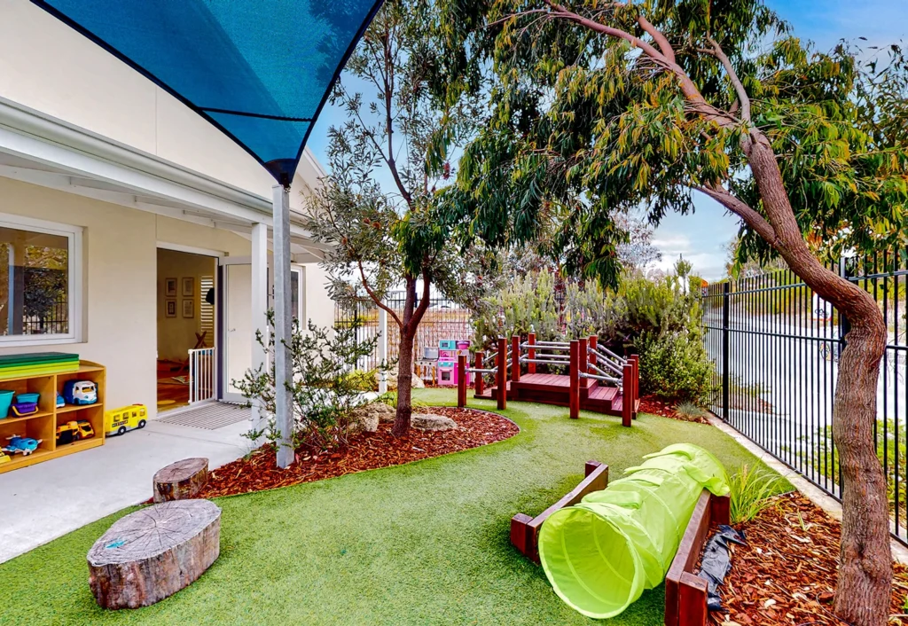 A kindergarten outdoor play area with artificial grass, a play tunnel, and wooden structures. Trees and a fence line the perimeter, and a canopy provides shade near the building.