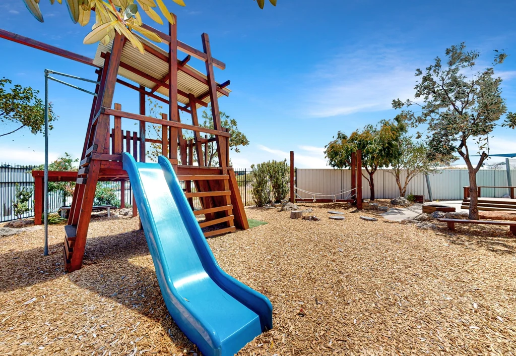 A children's playground at the day care features a wooden climbing structure, a blue slide, and wood chip ground cover. Tree branches are visible in the foreground, with a blue sky in the background.