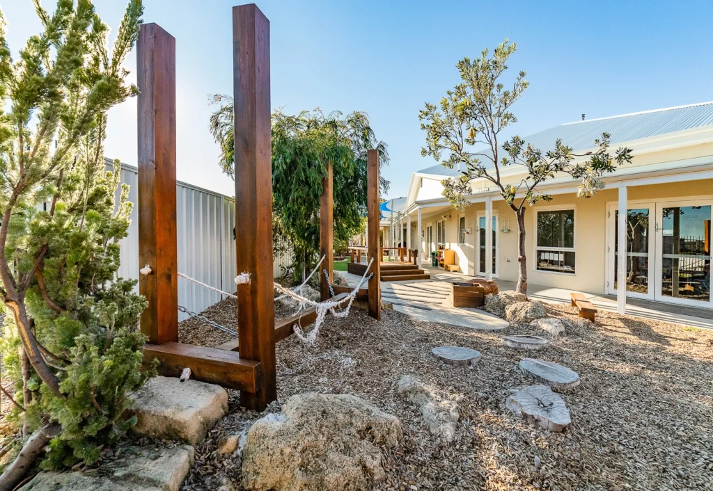 Outdoor area with wooden climbing structures, trees, and rocks, ideal for a day care. A modern building stands in the background. The ground is covered with mulch, and stepping stones create a playful path leading to the building.