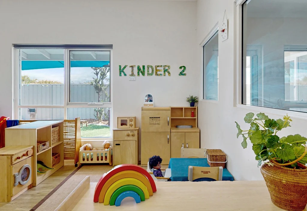 A bright, organized preschool classroom labeled "Kinder 2" with wooden toys, a kitchenette play area, and colorful decor, including a rainbow stacker in the foreground. Large windows allow natural light to fill this inviting day care space.