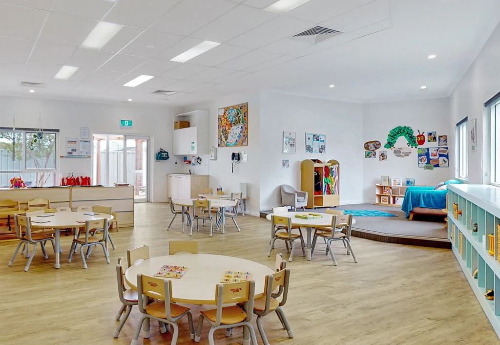 A brightly lit classroom designed for early learning, featuring round tables and chairs, colorful posters on the walls, shelves filled with books and toys, and a cozy reading nook with a sofa and carpet.