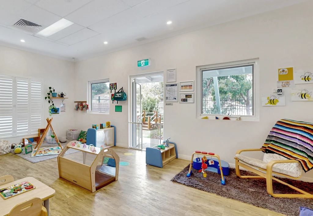 A bright, well-organized preschool room with various toys, learning stations, and art on the walls. Large windows provide natural light and colorful accents add a cheerful atmosphere, fostering an ideal early learning environment.