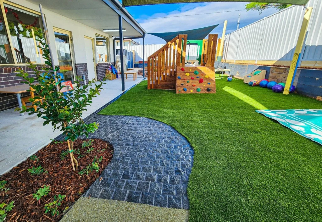 Outdoor play area with artificial turf, a wooden climbing structure, a small tree, and various toys. Pathway leads from patio to play area surrounded by a fence.