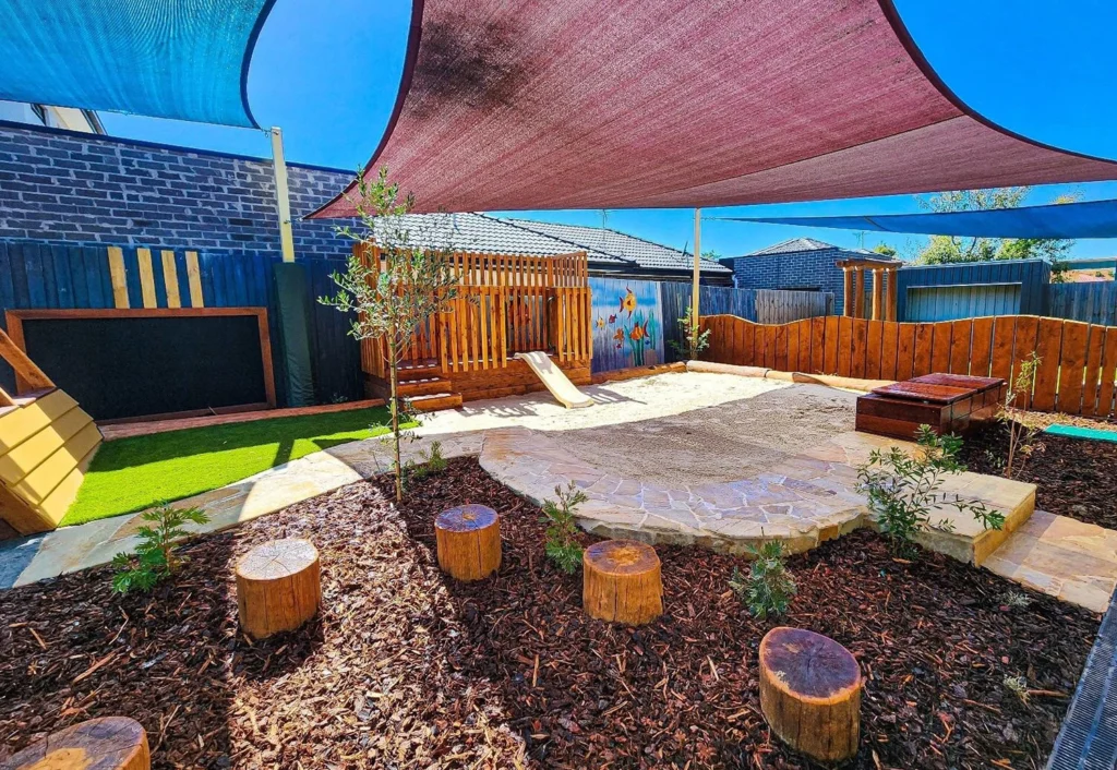 Outdoor play area with wooden stumps, a sandbox, a slide, a small stage, and shade sails overhead. The area is surrounded by a wooden fence and has a mixture of mulch and stone surfaces.