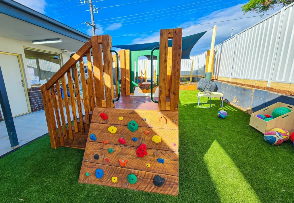 A small outdoor playground with a wooden climbing wall, grassy surface, various balls, a bench, a large box of toys, and shade sails. A building is on the left side.