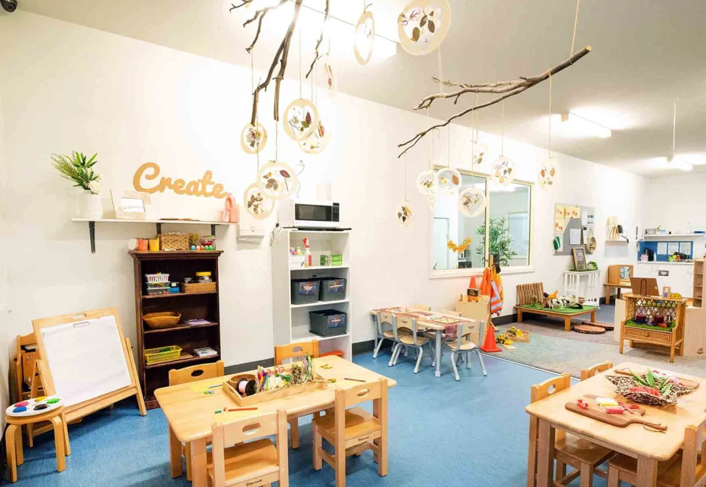 A brightly lit classroom with small wooden tables and chairs, educational toys, art supplies, a whiteboard easel, and decor emphasizing a creative and interactive learning environment.