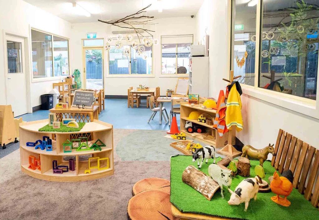 A colorful, well-organized classroom with various educational toys and activity areas, including models of animals, blocks, and craft supplies. The room has large windows and child-sized furniture.