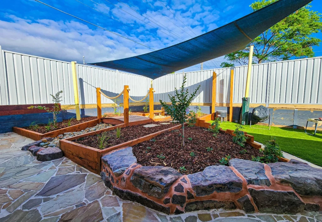 A backyard garden with raised planters and mulch, shaded by a large cloth canopy. The ground is a mix of stone and grass, and a fence lines the perimeter.