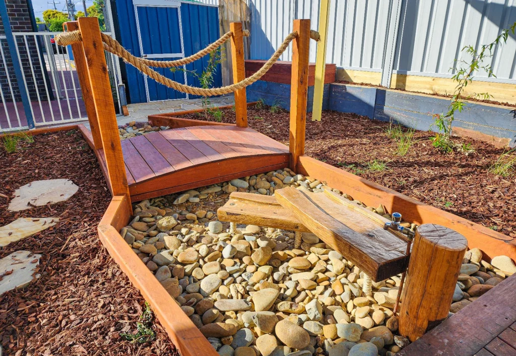 A small wooden bridge over a dry stone bed with a water play feature in a fenced outdoor area.