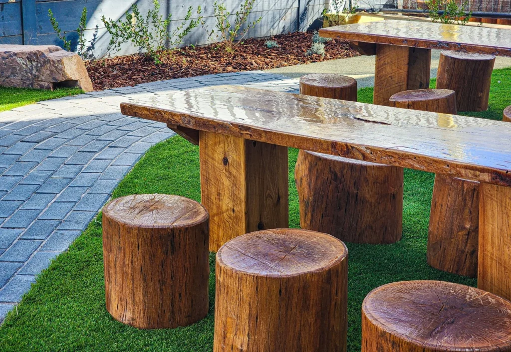 Outdoor seating area with wooden tables and cylindrical stools on a grassy surface, adjacent to a curved stone-paved walkway and landscaped garden.