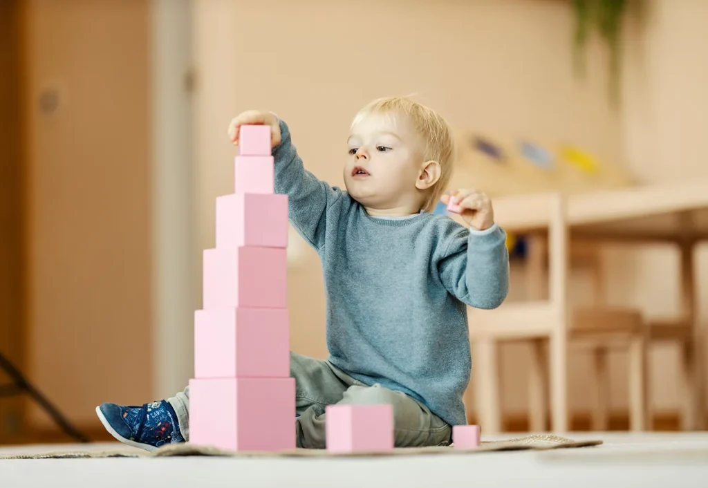 A toddler in a blue sweater is sitting on the floor, stacking pink blocks to form a tall tower.