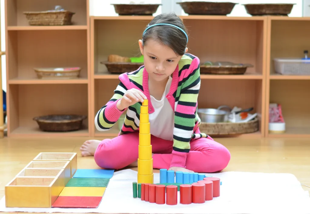 A child, dressed in a striped sweater and pink pants, builds a tower with colorful cylindrical blocks in a classroom setting.