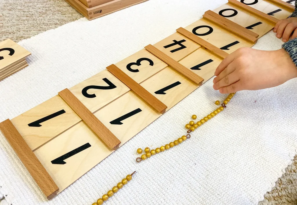 A child's hand is arranging yellow beads and wooden number tiles in sequence on a white mat, likely part of a Montessori math learning activity.