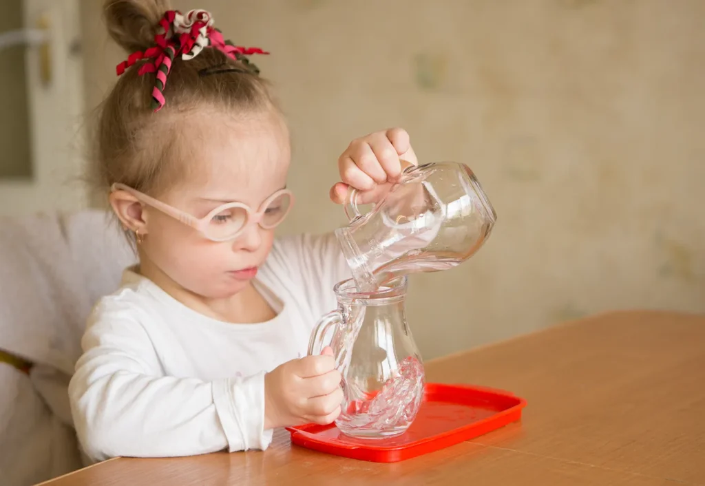 A young child with glasses carefully pours water from one clear pitcher to another while sitting at a table.