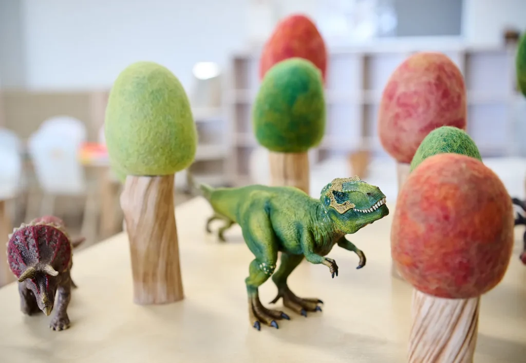 Toy dinosaurs are positioned among felt and wooden trees on a light-colored table.