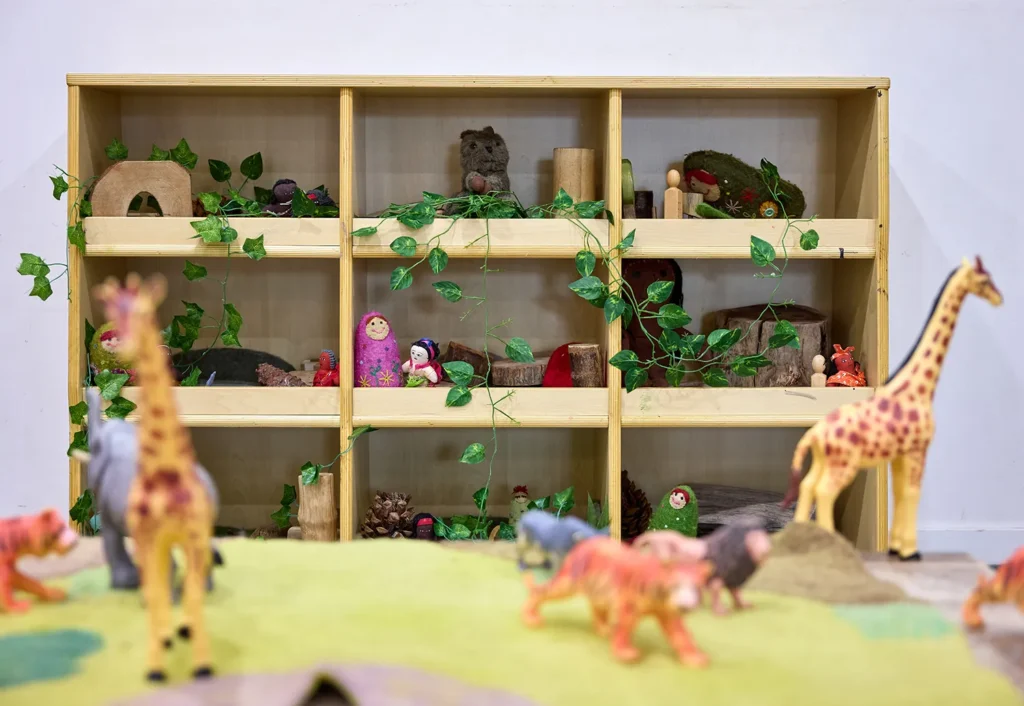 A toy zoo set on a table in a childcare room with a shelving unit filled with natural wooden toys.