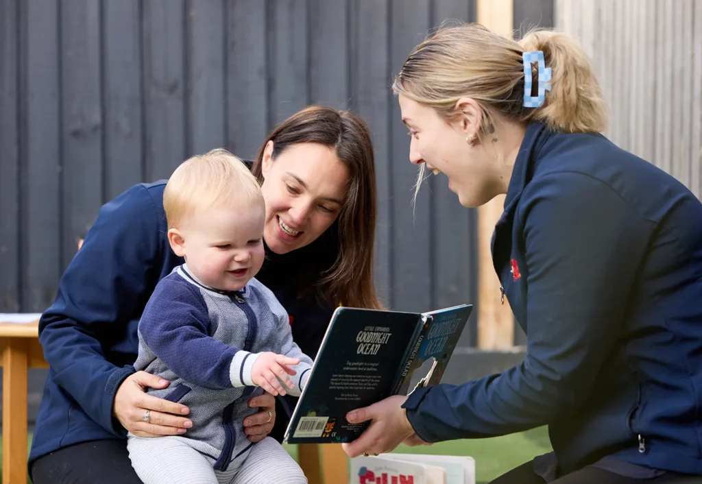 Two early learning educators and a baby looking with excitement at a book.