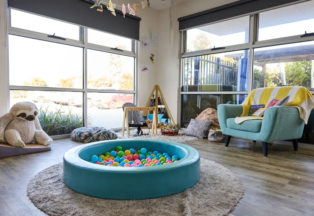 A cozy indoor playroom designed for early learning, featuring a ball pit, an armchair with a throw blanket, a stuffed sloth, various toys, and large windows with a view of the outdoors.