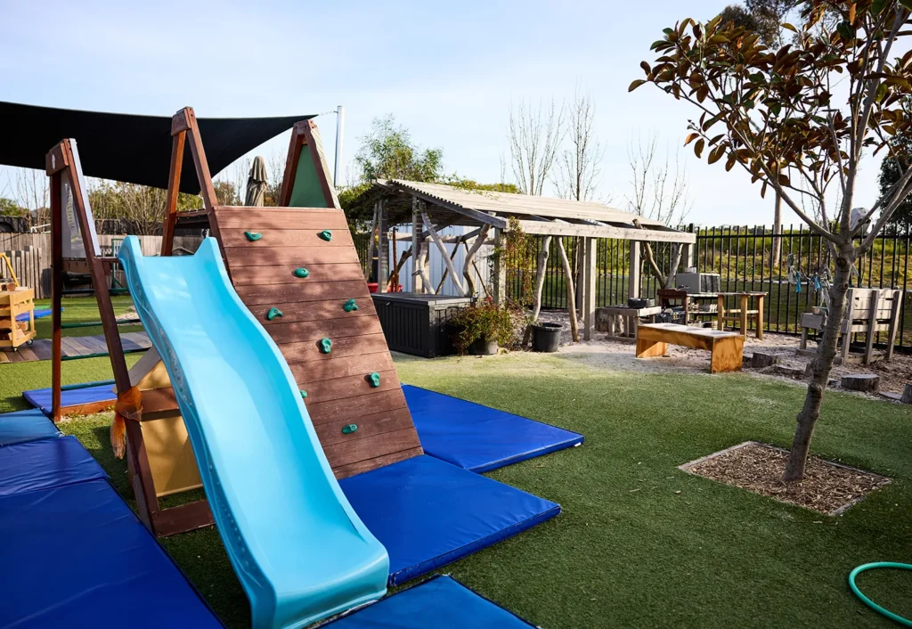 A playground area designed for preschoolers, featuring a blue slide attached to a wooden climbing structure. The ground is covered with blue mats and artificial grass. There's a tree and a backdrop of a fenced area with a wooden shed, perfect for day care fun.