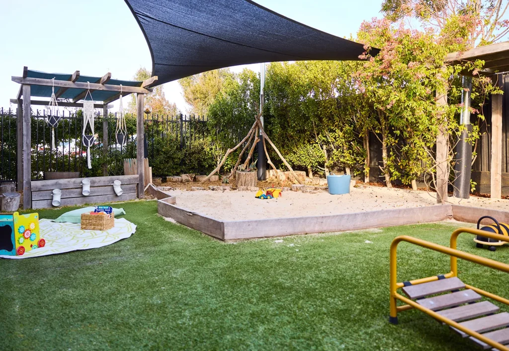 A delightful children's outdoor play area at the preschool features a sandbox with toys, shaded by a canopy. There are various play structures, a grassy area, and surrounding trees and bushes, creating an ideal environment for early learning and fun.