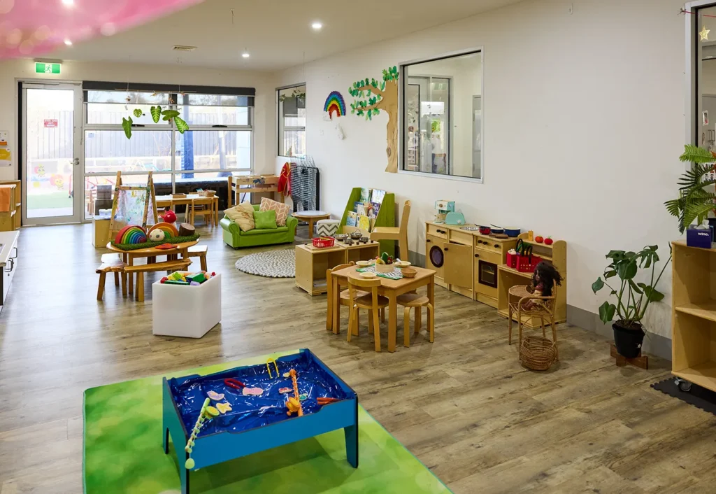 A preschool classroom designed for early learning, featuring various play areas including a water play table, a reading nook, and a kitchen set. The room is well-lit with natural light and decorated with children's artwork.