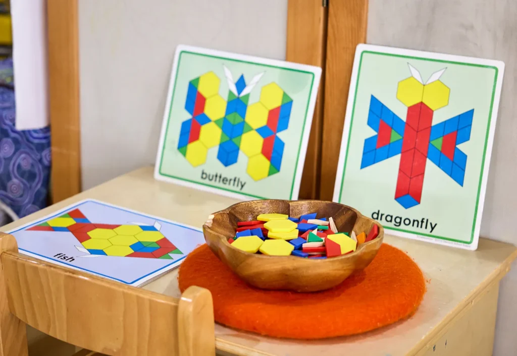 Colorful geometric shapes in a wooden bowl are on a table alongside pattern cards labeled "butterfly," "dragonfly," and "fish" for creating corresponding insects using the shapes, perfect for engaging preschool or kindergarten activities.