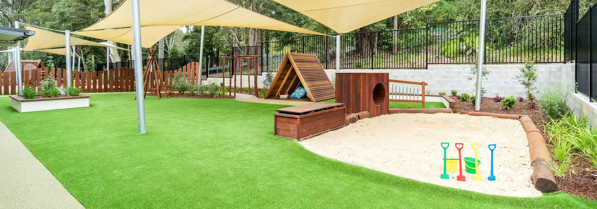 Outdoor playground at Central Coast child care centre featuring a sandpit with toy buckets and shovels, shade sails and wooden play structures.