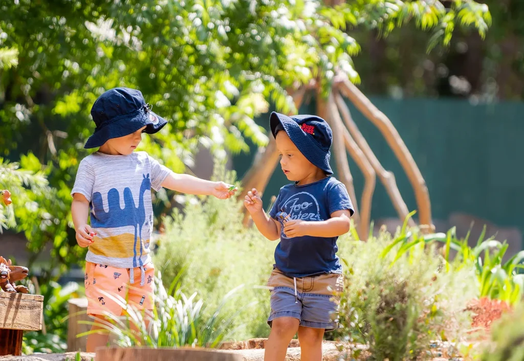 Two young children wearing Busy Bees branded sun hats playing in a garden at Follyfoot Farm Juniors, with one child handing something to the other.