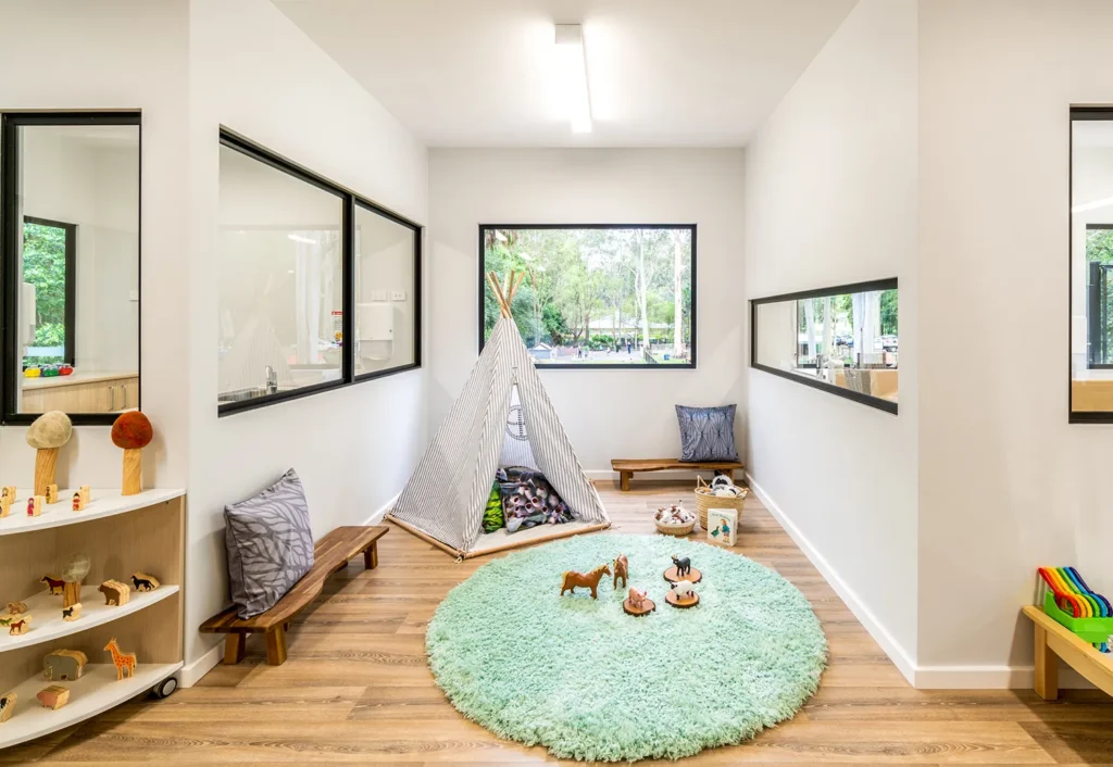 A bright, neatly arranged playroom with a soft round green rug, toy animals, and a fabric teepee tent. Large windows provide stunning outdoor views of the Central Coast day care's playground.