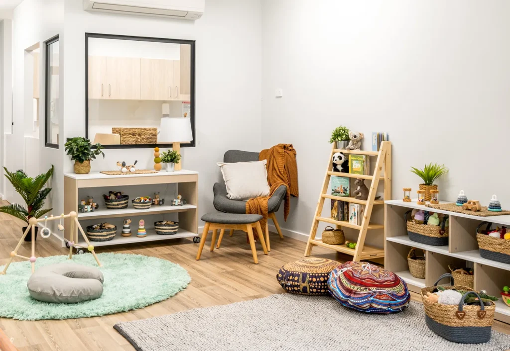 A cozy nursery day care room with various wooden toys on shelves, a cushioned chair, decorative pillows on the floor, and a soft green rug.