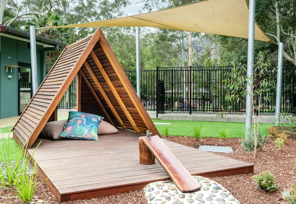 The outdoor play area at Central Coast childcare centre with a wooden a-frame cubby house filled with cushions, and a water play feature and tap.