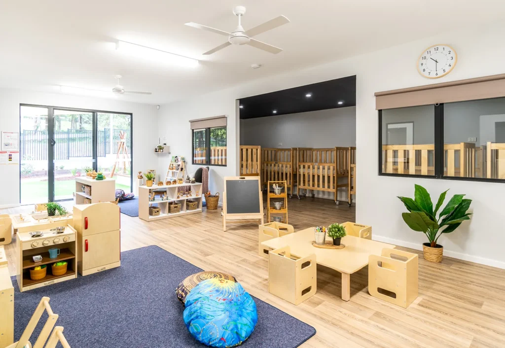 A brightly lit nursery day care room with wooden furniture, small tables and chairs, a cozy corner with cushions, and an adjoining cot room.