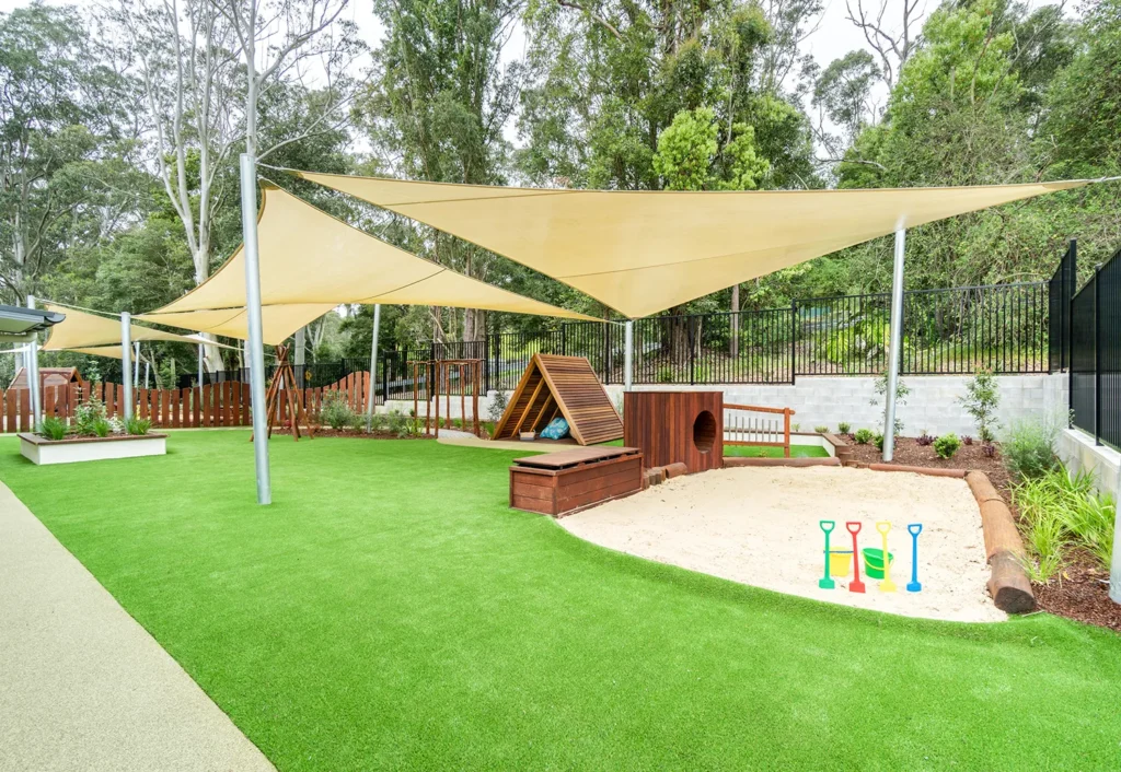 Outdoor playground at Central Coast day care featuring a sandpit with toy buckets and shovels, shade sails and wooden play structures.